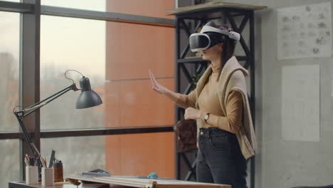 Modern-woman-engineer-designer-in-the-office-with-large-windows-stands-in-a-virtual-reality-helmet-uses-gestures-to-manage-the-project-without-leaving-the-office.-Construction-control.-Design-project-of-the-building-and-interior.